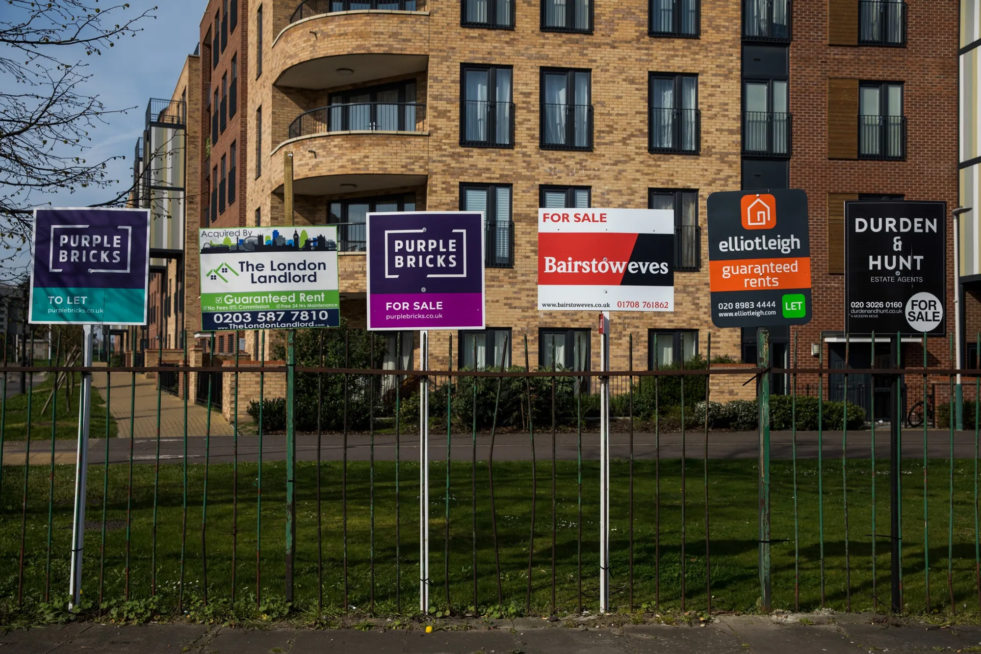A row of estate agents signs are attached to railings next to a block of residential apartments in Romford, U.K.