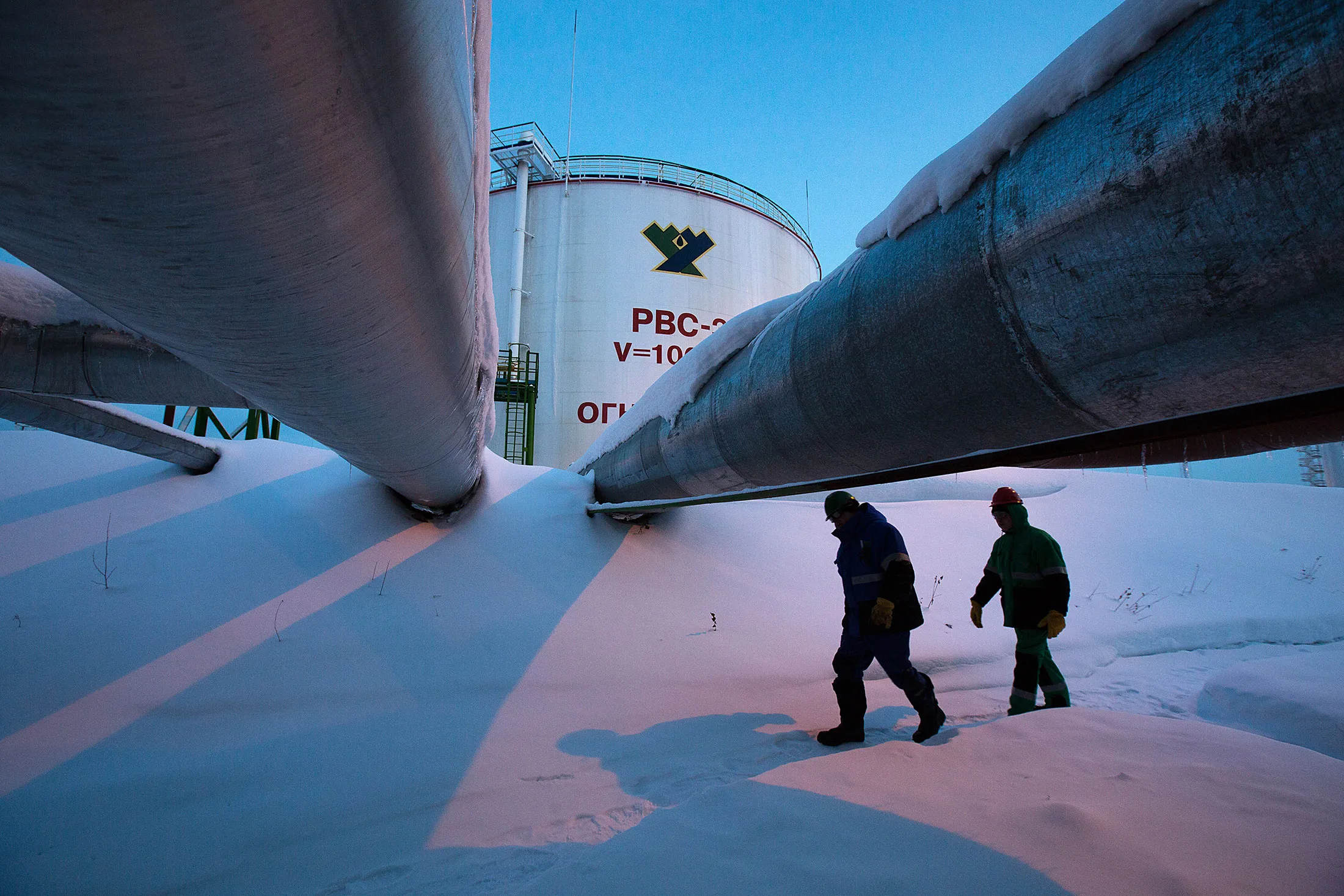 Employees pass beneath pipes leading to oil storage tanks at the central processing plant for oil and gas at the Salym Petroleum Development oil fields near the Bazhenov shale formation in Salym, Russia, on Tuesday, Feb. 4, 2014.
