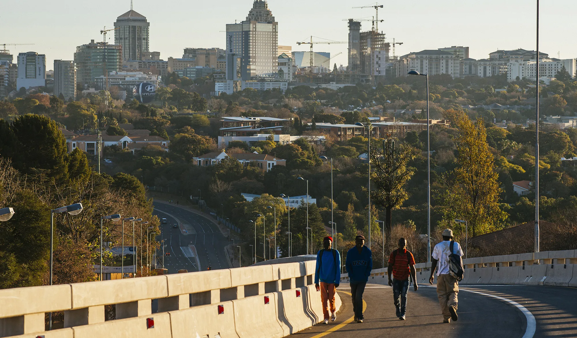 Pedestrians walk along a roadway back-dropped by commercial and residential buildings in the Sandton district of Johannesburg, South Africa.