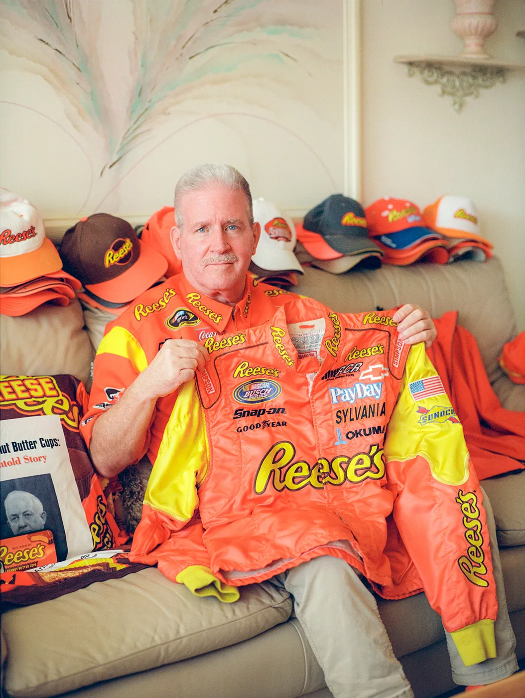 Brad Reese sitting on a sofa amongst several branded Reese's items including a leather jacket, throw pillow, over 10 hats and other apparel items.