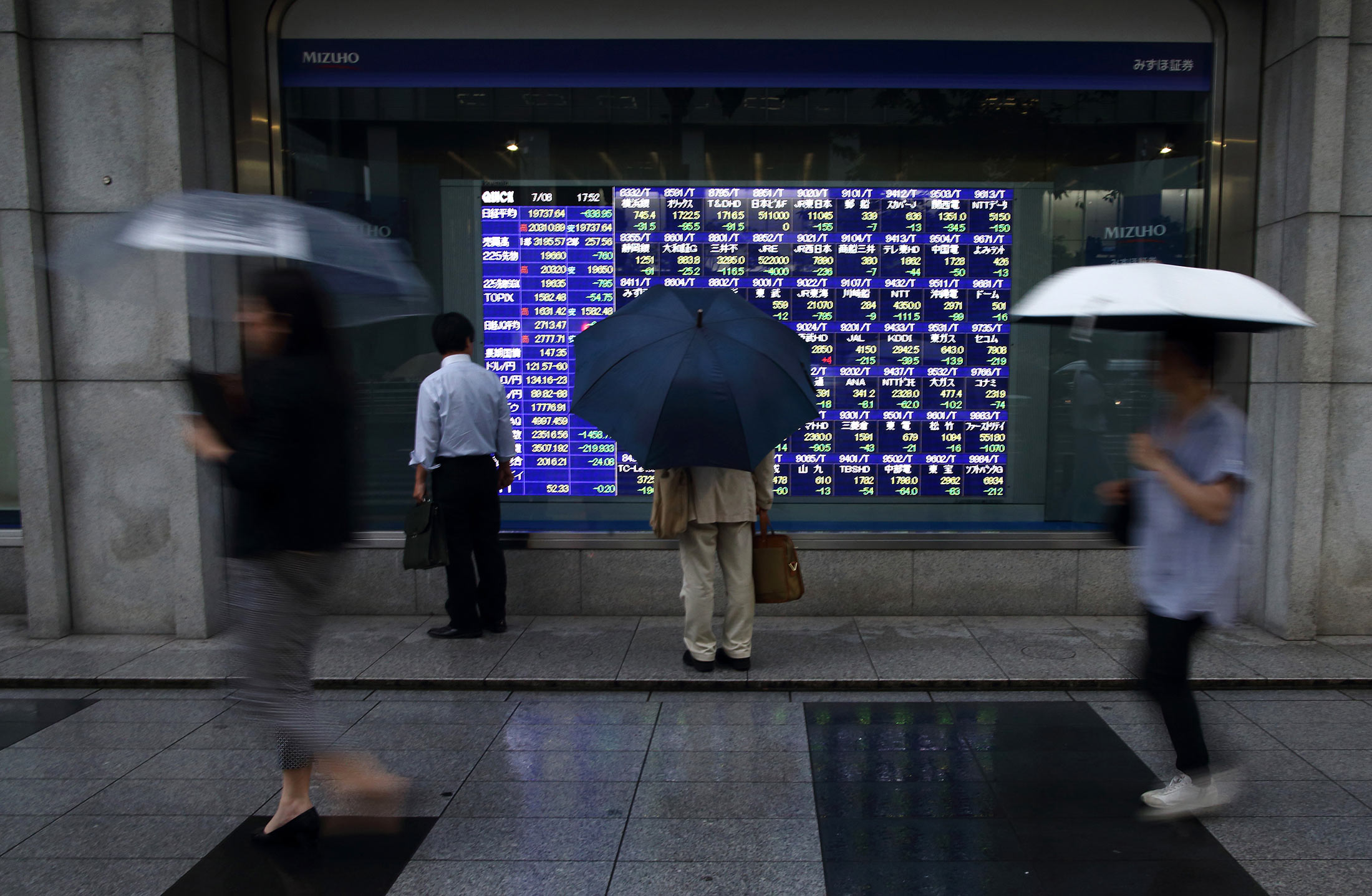 Pedestrians holding umbrellas walk past an electronic stock board outside a securities firm in Tokyo, Japan, on Wednesday, July 8, 2015. Japanese stocks fell, with the Topix index dropping by the most in more than a year, amid concern that ChinaÕs equity rout is spreading.
