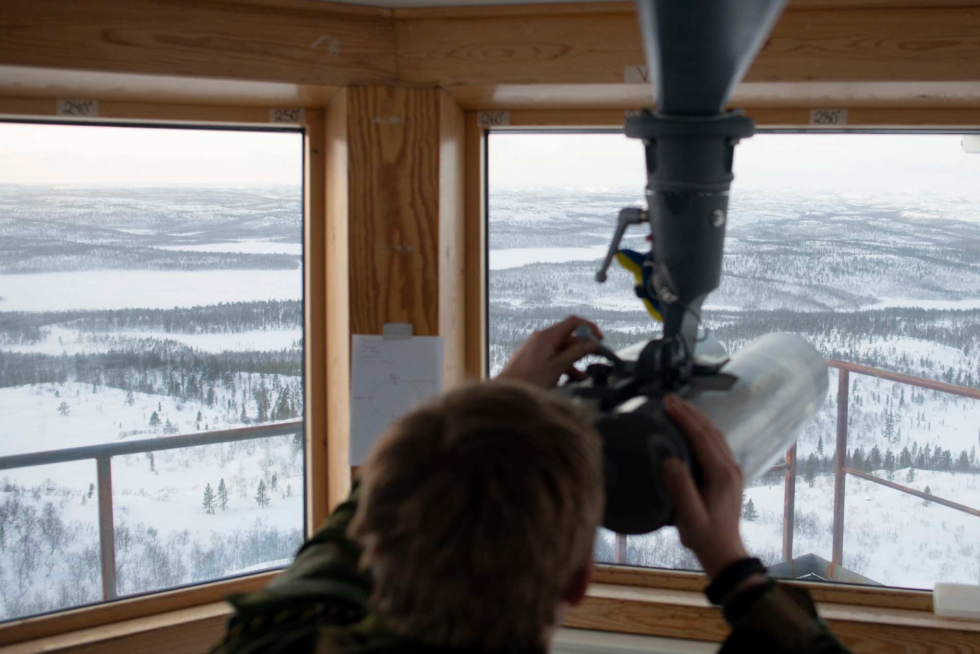 A soldier, the back of their head and arms visible, looks through a telescope attached to the ceiling. It looks through the window of a room in a high-vantage point. The landscape beyond is an Arctic wilderness of snow-covered land and forests stretching away to the horizon.