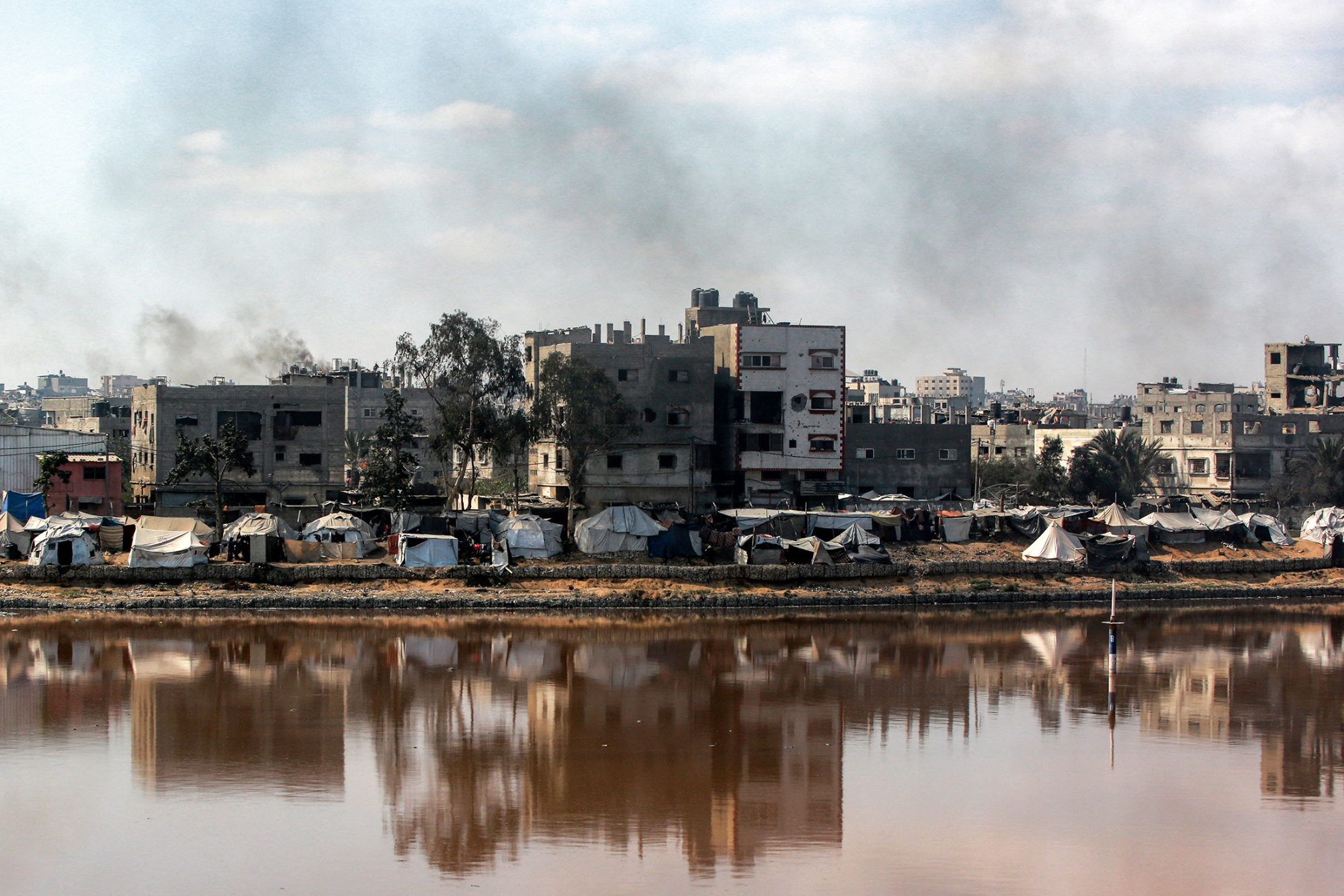 A photograph of a stark, urban landscape. In the foreground is a body of still, murky water, depicting a wastewater collection pond in Gaza City. On the opposite bank, a dense line of makeshift tents and shelters is crowded together. Directly behind this camp, several multi-story concrete buildings, some of which appear damaged, rise into a hazy blue sky. Plumes of gray smoke are visible rising from behind the city buildings.