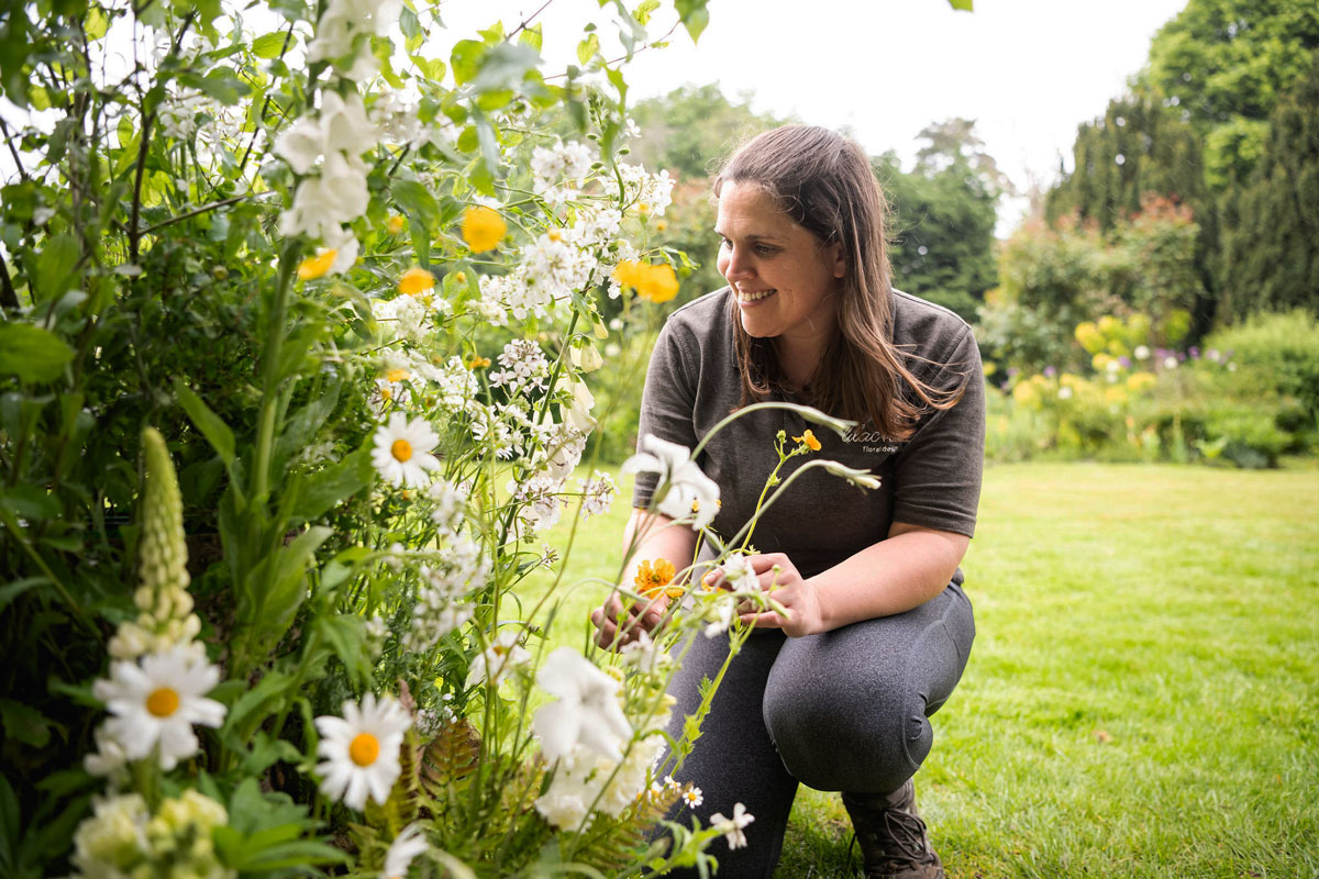Florist Rebecca Yussuf is squatting on a lush green lawn before a bed of daisies, foxgloves and other pretty yellow and white flowers.