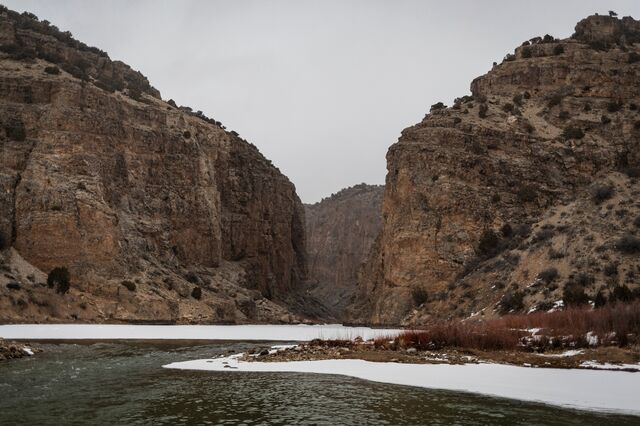 A photo of a river with icy and snowy banks flowing in the foreground, and steep red, cliffs along the river in the background.