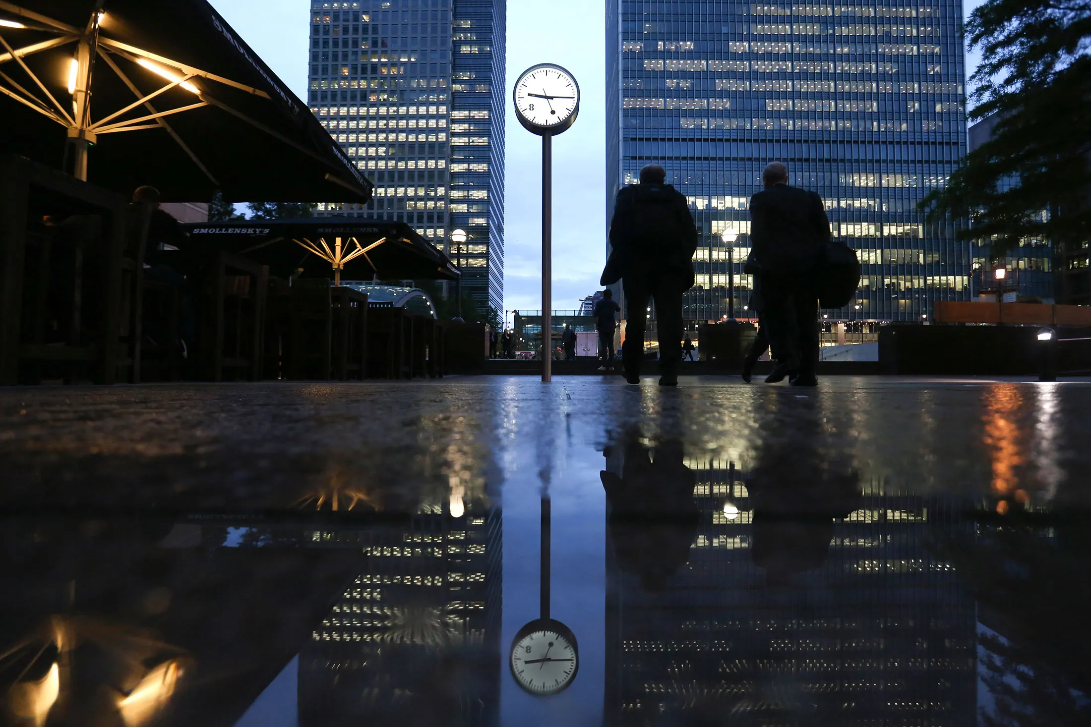 Commuters walk past buildings illuminated at night in the Canary Wharf business, financial and shopping district of London, U.K., on Oct. 28, 2015.
