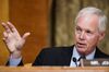 Senator Ron Johnson is speaking in front of a senate committee. It’s a close-up portrait of him, he’s framed above the chest, and is looking to the left of the picture, his hand is gesturing in that direction as well. He has grey hair and is wearing a dark suit with a light blue shirt.