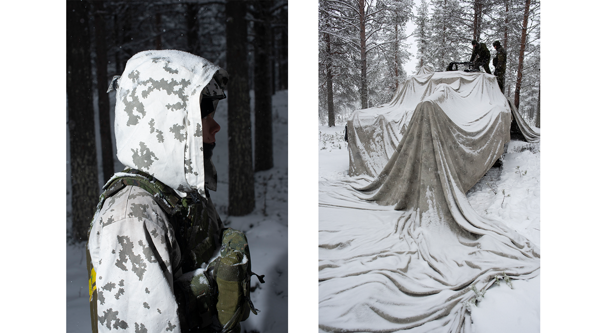 On the right, the shape of a tank with the muzzle pointed toward the camera can be discerned under a large white tarp. The tank sits in snow in the middle of an Arctic forest in Finland, with two soldiers wearing green uniforms standing on top of it. On the left, the upper body and head of a Finnish soldier is framed close up in profile. They are wearing white camouflage, including a hood, which obscures their face. They are standing in snow amid tall trees.
