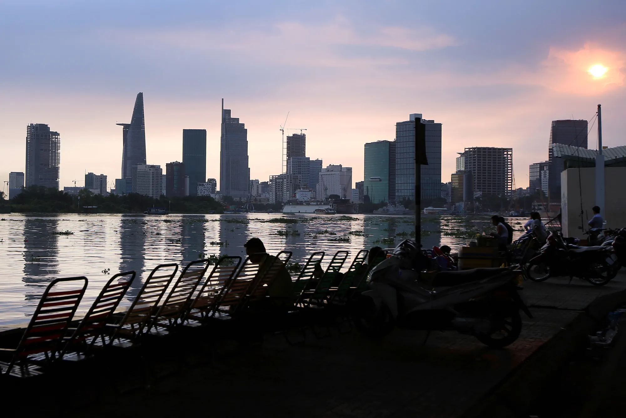 People sit along a waterfront as residential and commercial buildings stand in the background in Ho Chi Minh City, Vietnam.