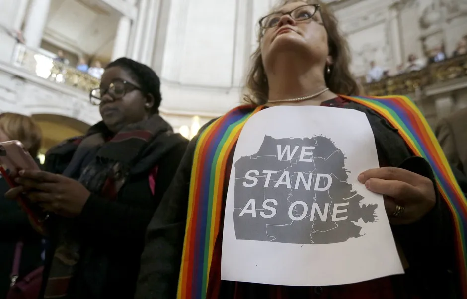 The Rev. Annie Steinberg-Behrman, right, provisional pastor with Metropolitan Community Church, holds a sign while listening to speakers at a meeting at City Hall in San Francisco by city leaders and community activists to reaffirm the city's commitment to being a sanctuary city in response to Donald Trump's support of deportations and other measures against immigrants Monday, Nov. 14, 2016. 