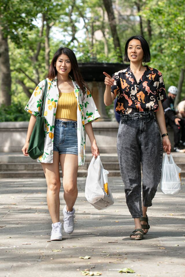 Yin Chang and Moonlyn Tsai walking in park. Yin Chang is wearing white shirt with flowers and fruits, denim shorts, yellow tank top, white sneakers carrying a plastic takeout bag. Moonlyn is wearing a shirt with animal pattern, grey jeans, sandals, is pointing at object, and carrying a plastic takeout bag.