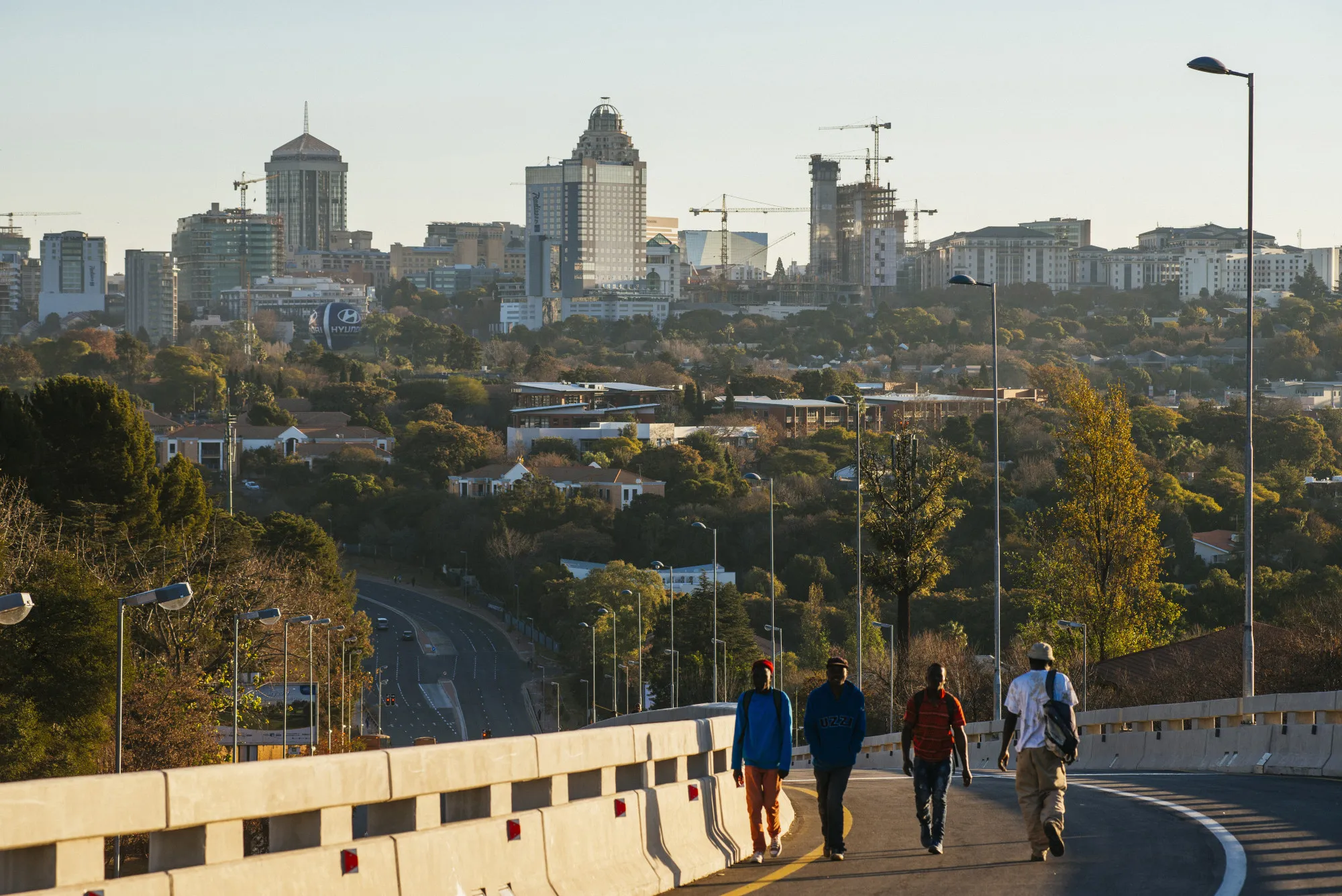 Pedestrians walk along a roadway back-dropped by commercial and residential buildings in the Sandton district of Johannesburg, South Africa.