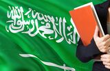 Learning Arabic language concept. Young woman standing with the Saudi Arabia flag in the background. Teacher holding books, orange blank book cover.