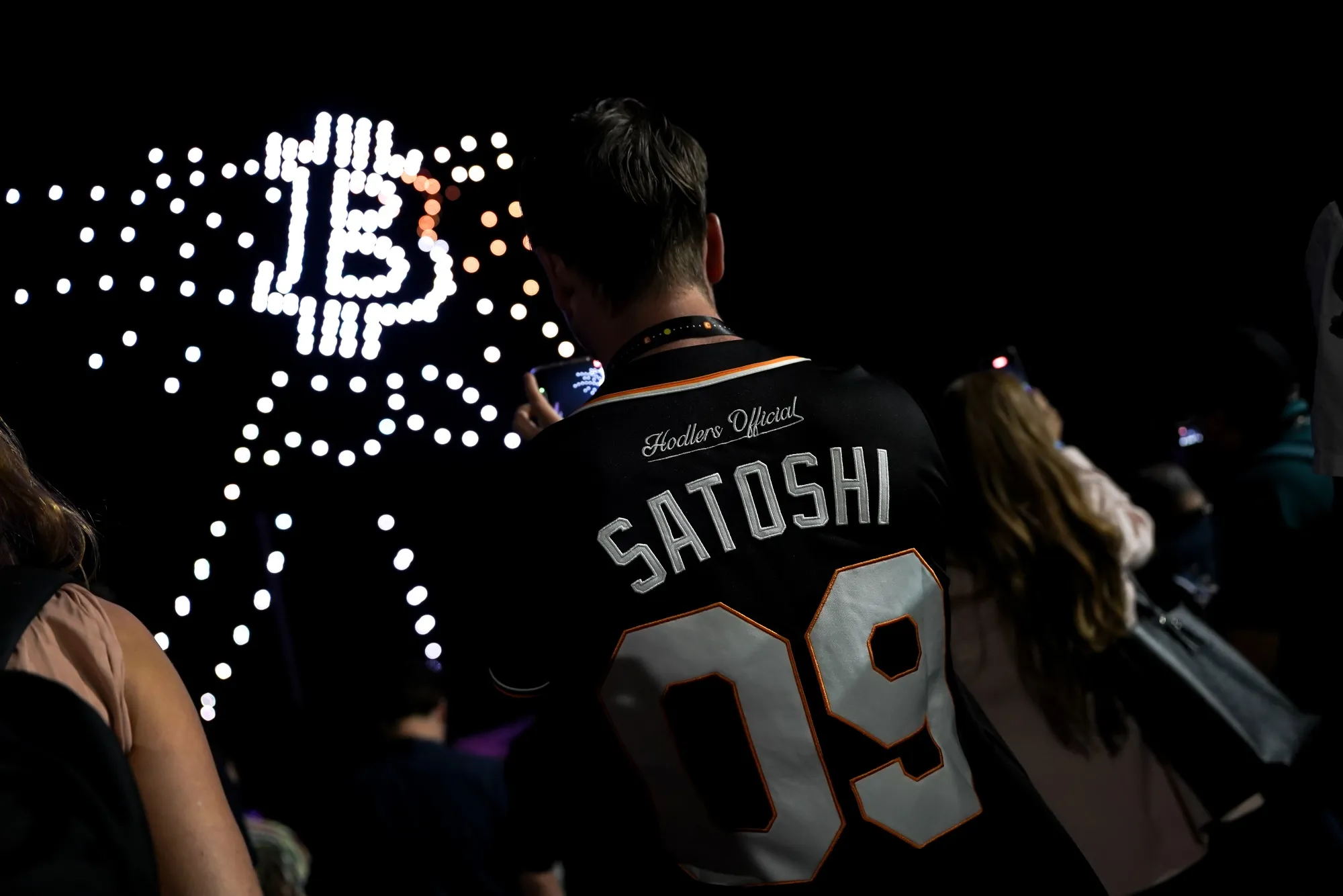 An attendee wears a Hodlers Official Team Bitcoin baseball jersey as drones form the shape of Bitcoin signag.