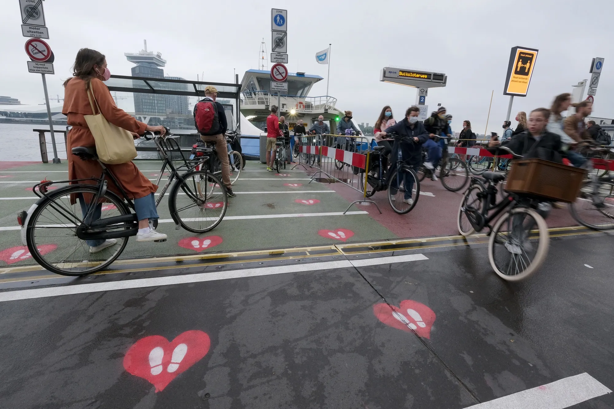 Commuters ride bicycles over heart-shaped social distancing signs at a IJ River passenger ferry dock near Amsterdam Central Station in Amsterdam.