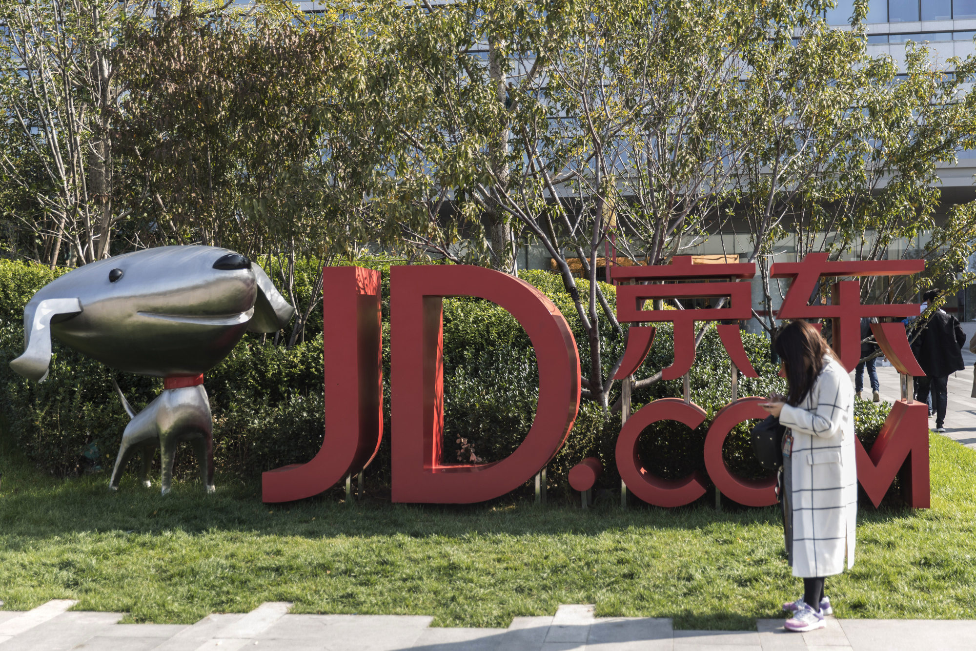 A woman stands next to signage incorporating the logo of JD.com Inc. and the company's mascot &quot;Joy&quot; at the company's headquarters in Beijing, China, on Monday, Oct. 23, 2017. JD.com is China's second-largest online mall.