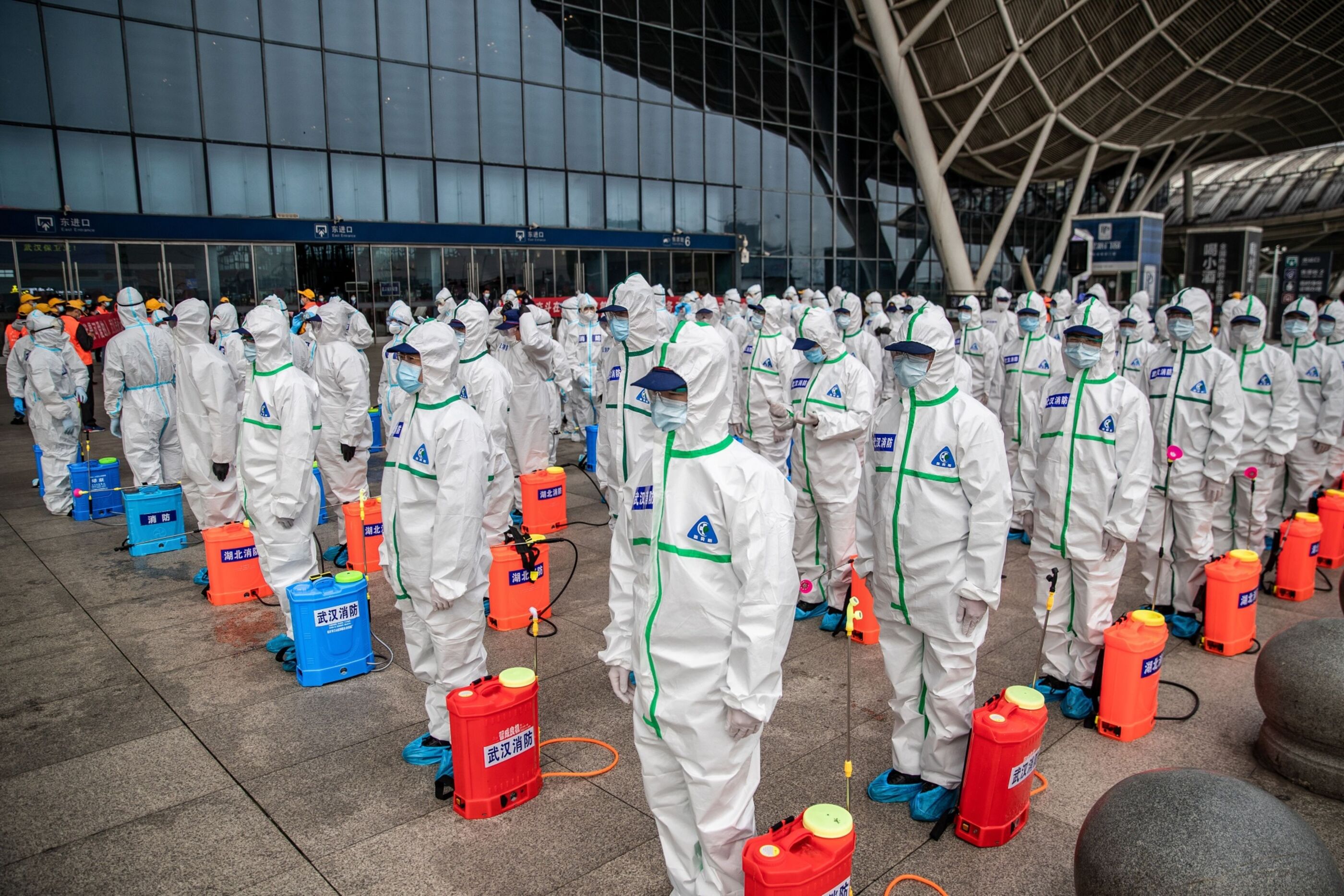 A group of people in white protective jumpsuits, face masks and goggles stand in rows, each with a tub of disinfectant and sprayer.