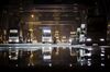 Trucks are reflected in a puddle as they wait in line to unload their containers onto CMA CGM SA's Benjamin Franklin container ship docked at the the Xiamen Songyu Container Terminal at night in Xiamen, China.