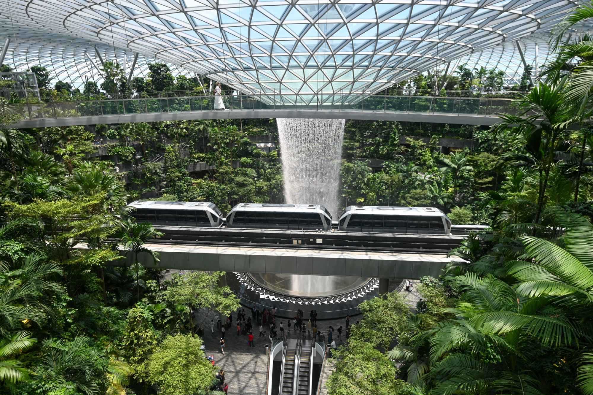 A woman (top) walks across the bridge next to the Rain Vortex, the world's tallest indoor waterfall, and a mono-rail train going through the Jewel Complex at Singapore Changi Airport in Singapore on April 10, 2025. Photographer: Roslan Rahman