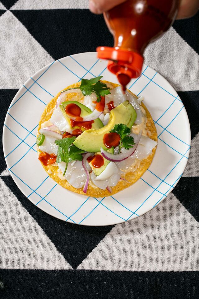 A tostada with avocado, hot sauce, cream, onion and cilantro on table with triangle patterned table cloth
