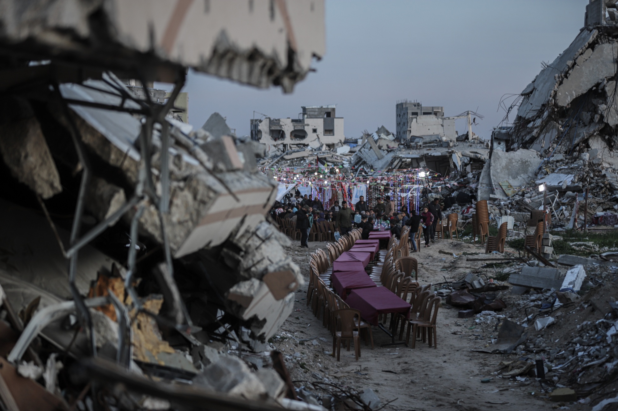 Palestinians attend an iftar dinner in the rubble of destroyed buildings. A row of tables with chairs on either side.