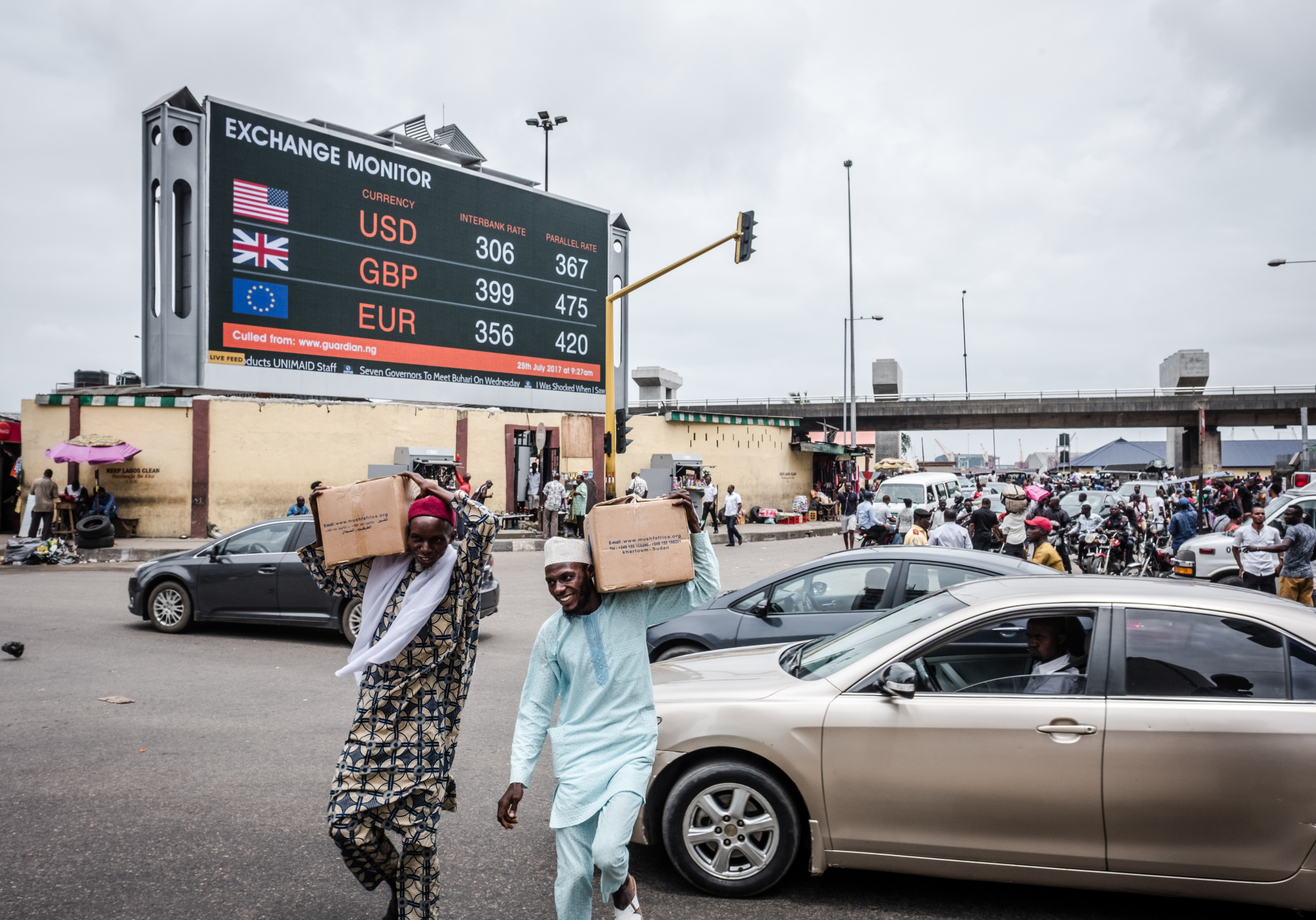 Pedestrians carry boxes of goods across a busy road near a giant advertising screen showing US dollar, British pound and euro foreign currency exchange rates in Lagos, Nigeria.