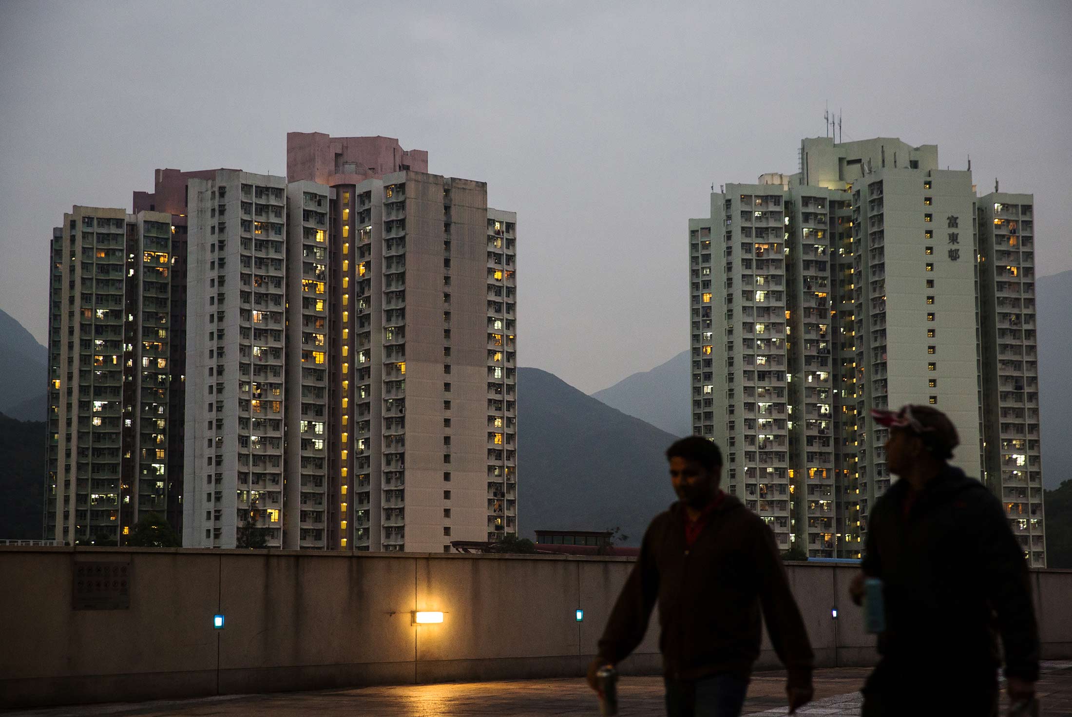 Pedestrians walk past residential buildings illuminated at night in the Tung Chung area of Hong Kong, China, on Wednesday, Feb. 3, 2016. Hong Kong is seeing negative-equity mortgages for the first time since September 2014.
