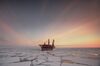 Picture of Russia’s offshore Prirazlomnaya oil platform sitting in the Pechora Sea in the Arctic region. The platform is a red industrial looking construction from which a flame bursts at the top. The landscape looks dark, it’s probably nightfall. The sea is partially covered with ice.