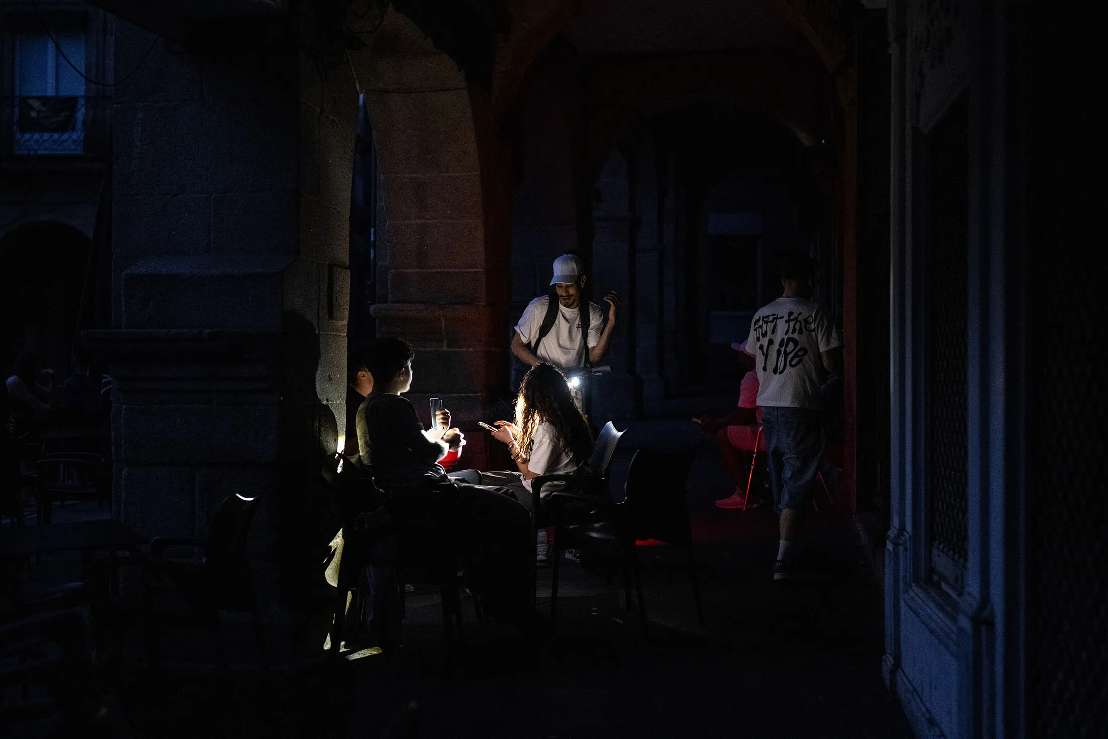 People sit at an outdoor café in Ourense, Spain, during a power outage on April 28, their faces illuminated by phone screens and flashlights in an otherwise dark arcade.