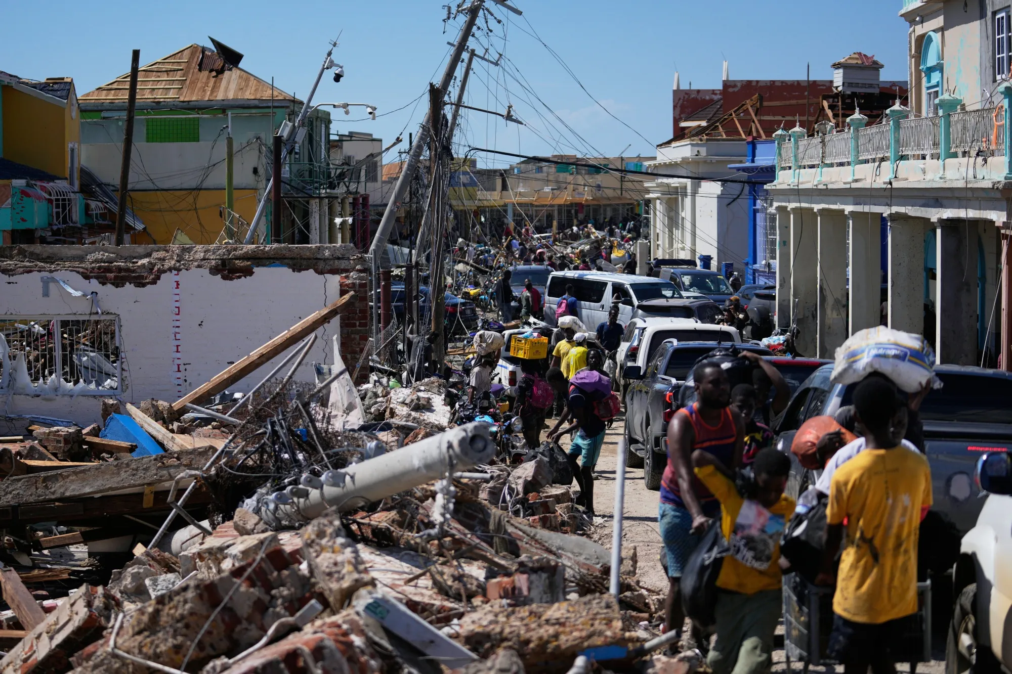 Street in a town lined with buildings that sustained heavy damage from high winds, with people walking through the ruins, some carrying bags or boxes over their heads