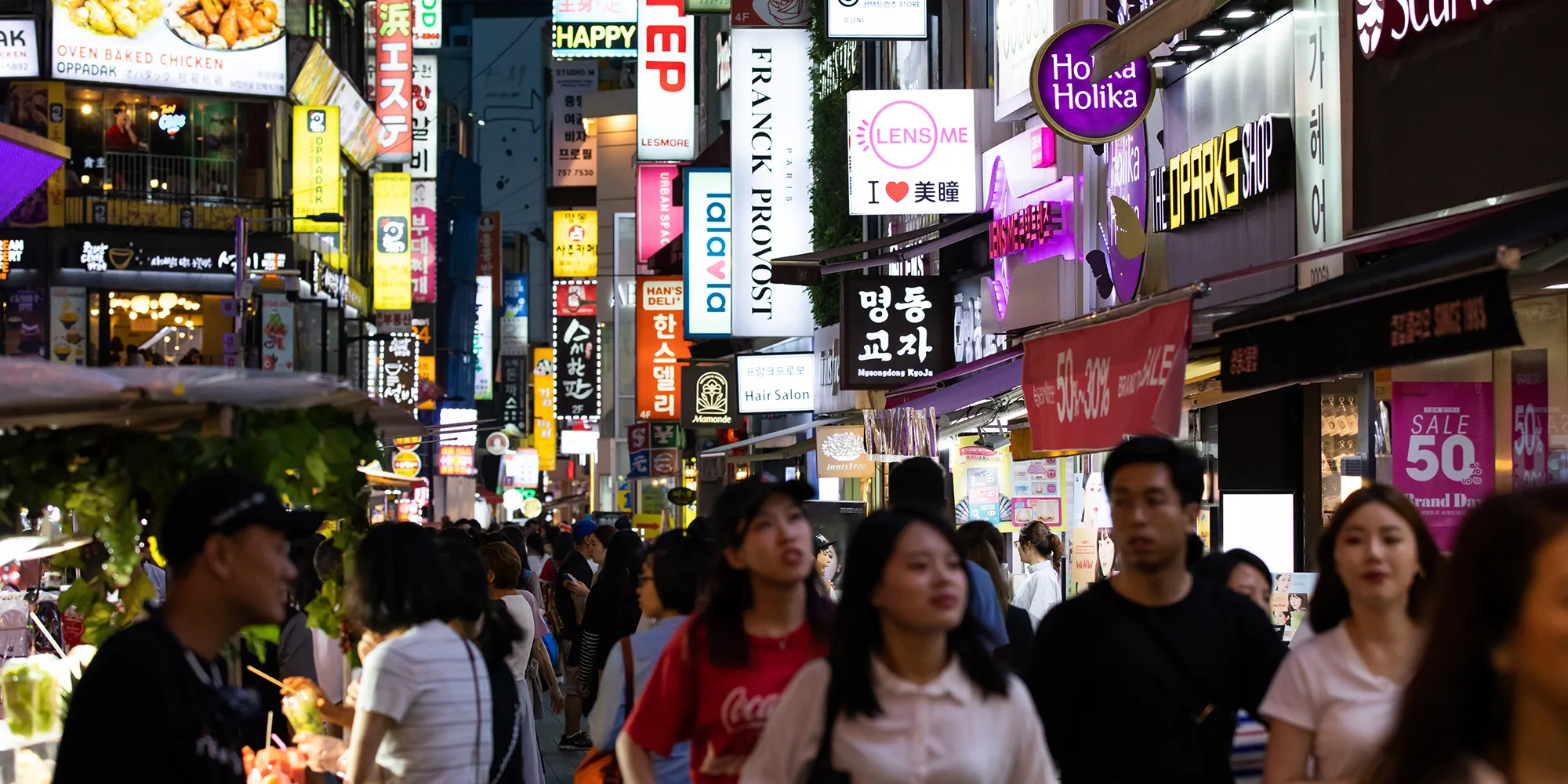 Pedestrians walk past illuminated store signs at night in the Myeongdong shopping district of Seoul.