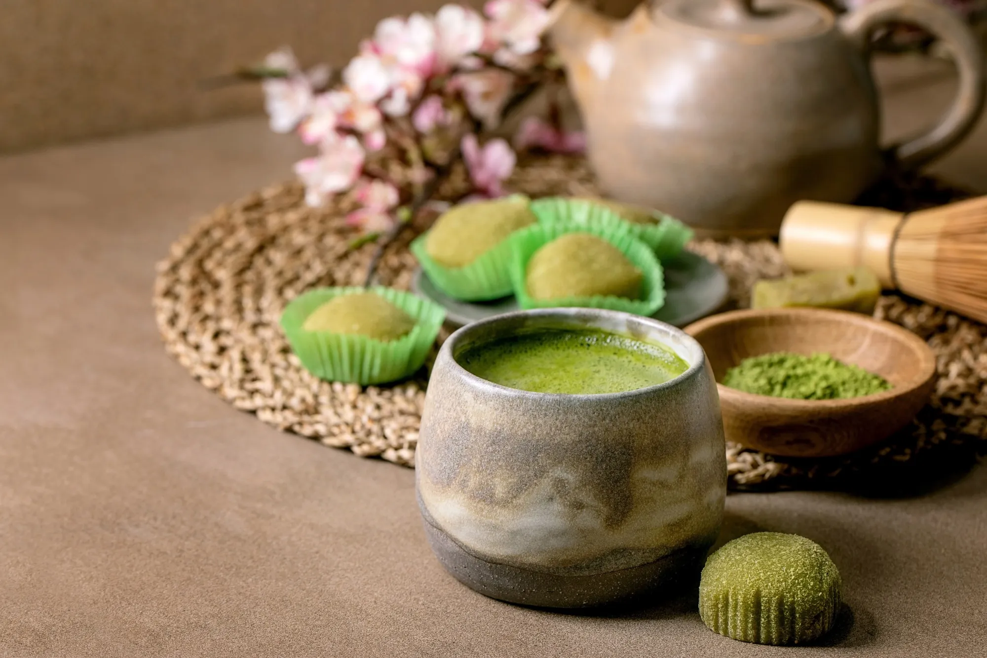 Asian rice dessert sweet green matcha mochi with cup of frothed matcha tea and green matcha powder in ceramic plate on brown table. Teapot and spring pink blooming branch behind. Copy space