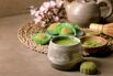 Asian rice dessert sweet green matcha mochi with cup of frothed matcha tea and green matcha powder in ceramic plate on brown table. Teapot and spring pink blooming branch behind. Copy space