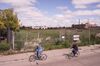 Two men riding bicycles past the fenced field, now with bushes removed