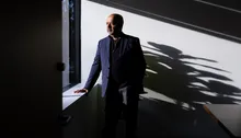 Haris Basit, wearing a blue blazer on top of a sweater stands looking out the window in a room with shadows on the wall to his left. The silhouette of a plant can be seen on the wall as the bright light from the window contours Basit's face. His right hand rests on the windowsill while his left is tucked into his pocket. 