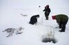 Three people in dark, thick winter coats and hoods are standing near a hole cut into the white ice and snow. Thin green rope extends into the water. There is a pile of fish to the side and one fish hooked on a line in mid-air.