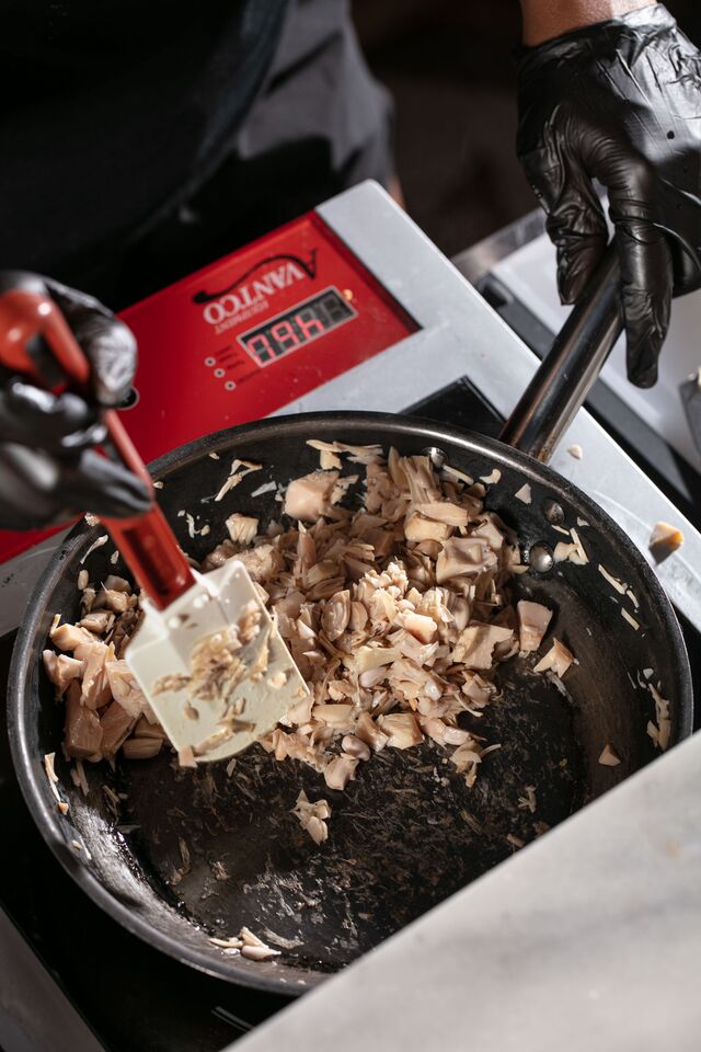 A pan with pieces of food being mixed. Cook is wearing black gloves and using a rubber spatula.