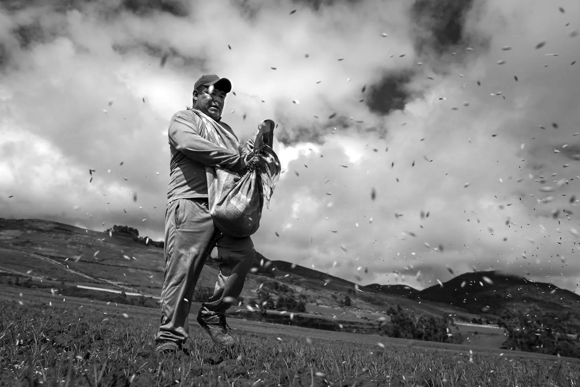 A Black and white image of a man reaching into a bag to scatter seeds in a field.