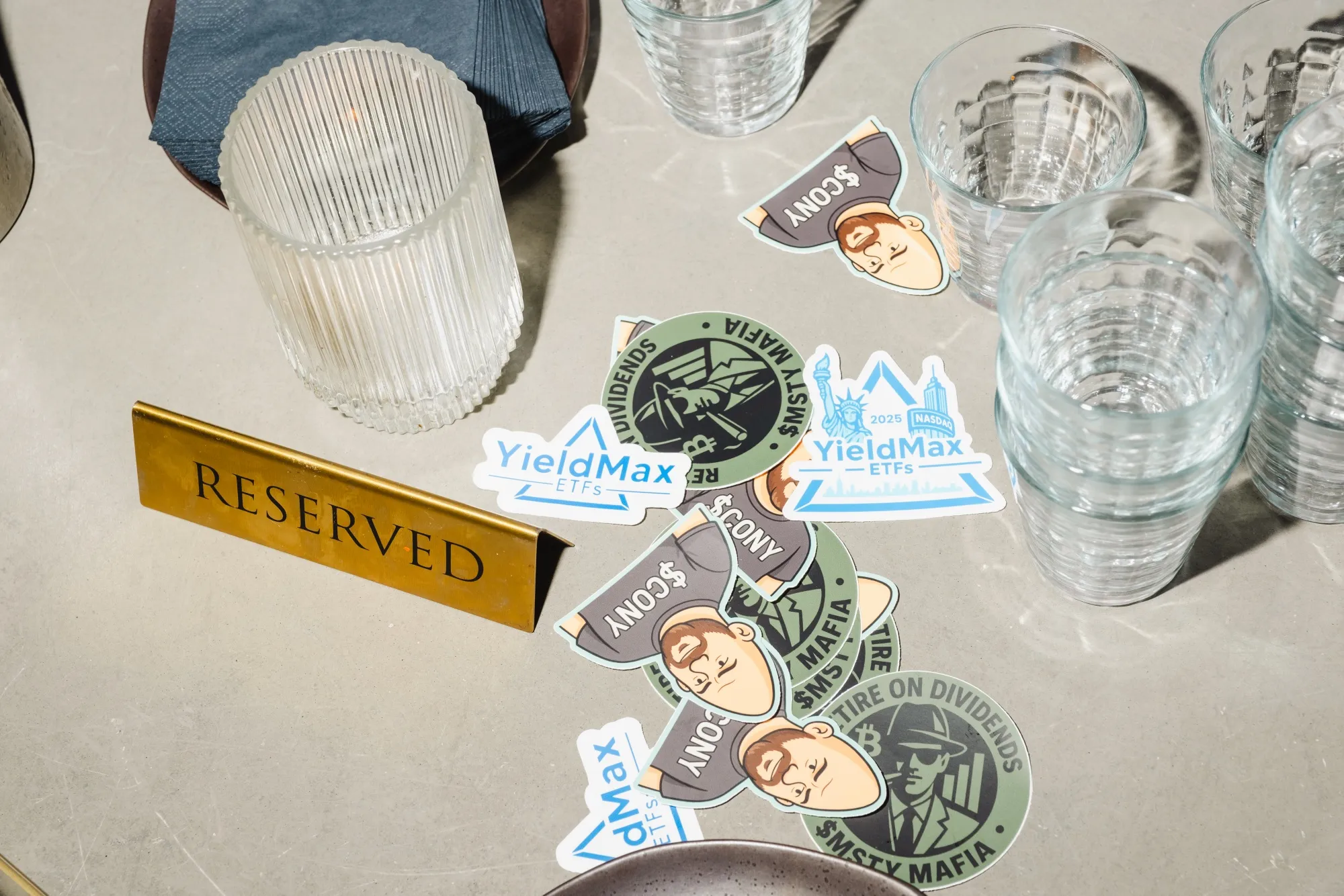 Stickers for CONY and other YieldMax ETFs lie on a table with empty glasses and a "reserved" place holder.