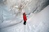 A picture of Putin standing in an ice cavern, looking past the photographer from afar. He’s wearing a red puffer, a red cap and black pants. He looks like he’s smiling.