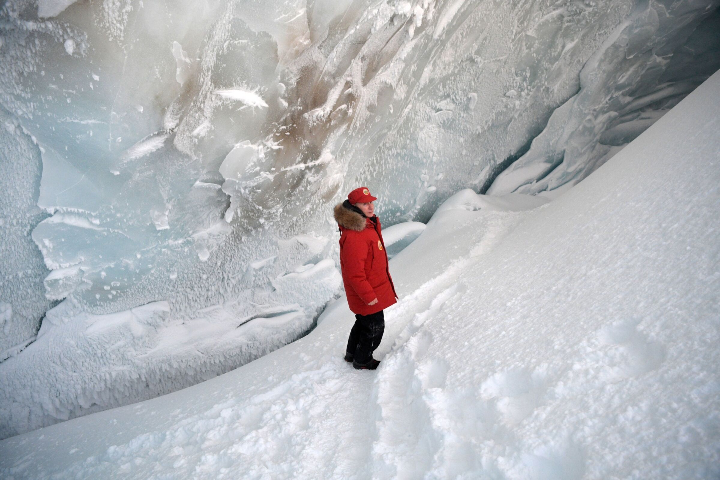 Putyin képe egy jégbarlangban állva, és messziről elnéz a fotós mellett.  Piros nadrágot, piros sapkát és fekete nadrágot visel.  Úgy néz ki, mintha mosolyogna.