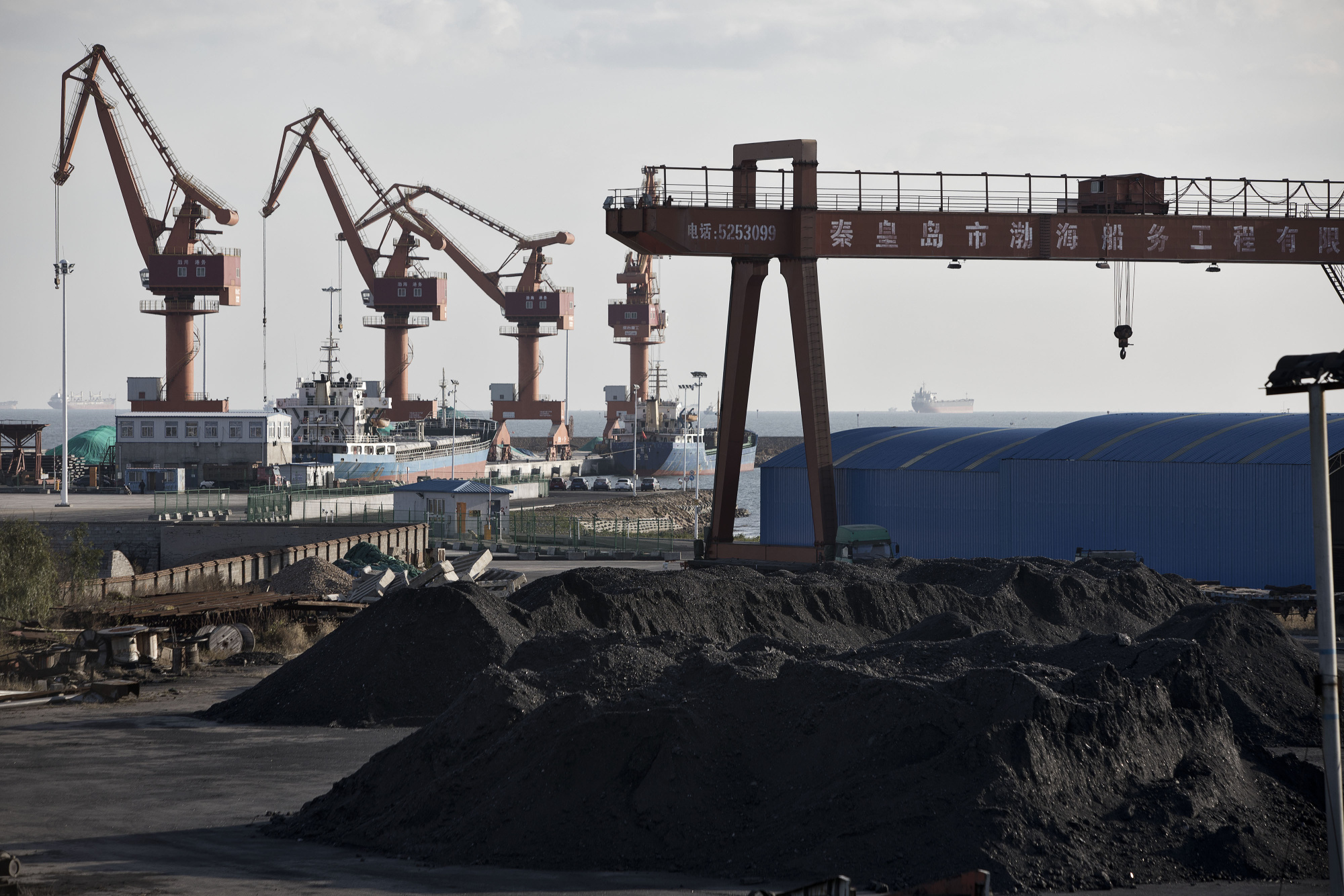 Piles of coal sit near port facilities as gantry cranes stand in the background at the Qinhuangdao Port in Qinhuangdao, China.