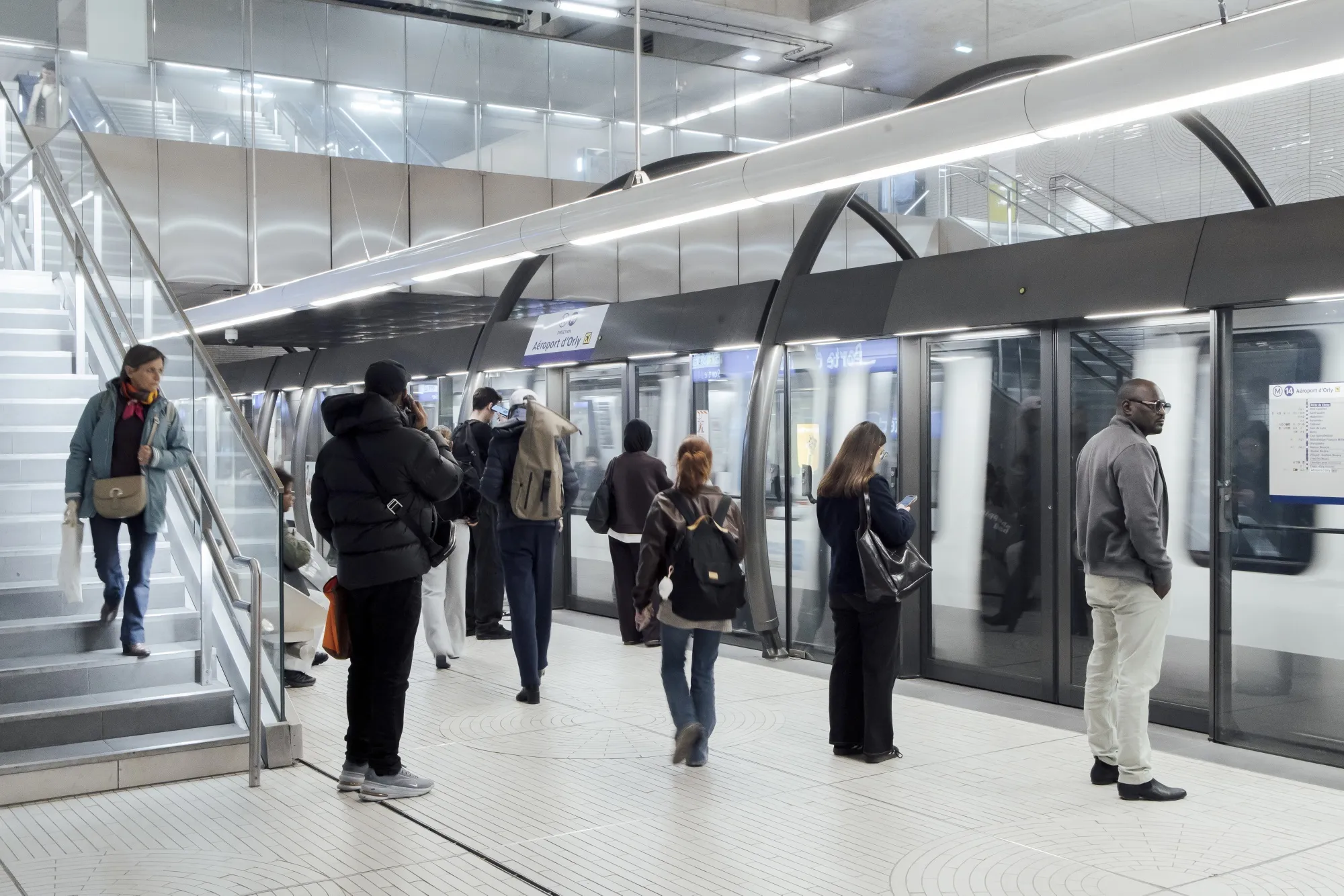A bright, modern subway station platform with a sleek, minimalist design. The station features glass safety barriers with automatic sliding doors separating the platform from the tracks. Several commuters are scattered across the platform. Some are walking toward the train doors, one person is descending the stairs, and others are standing still, waiting for the train.