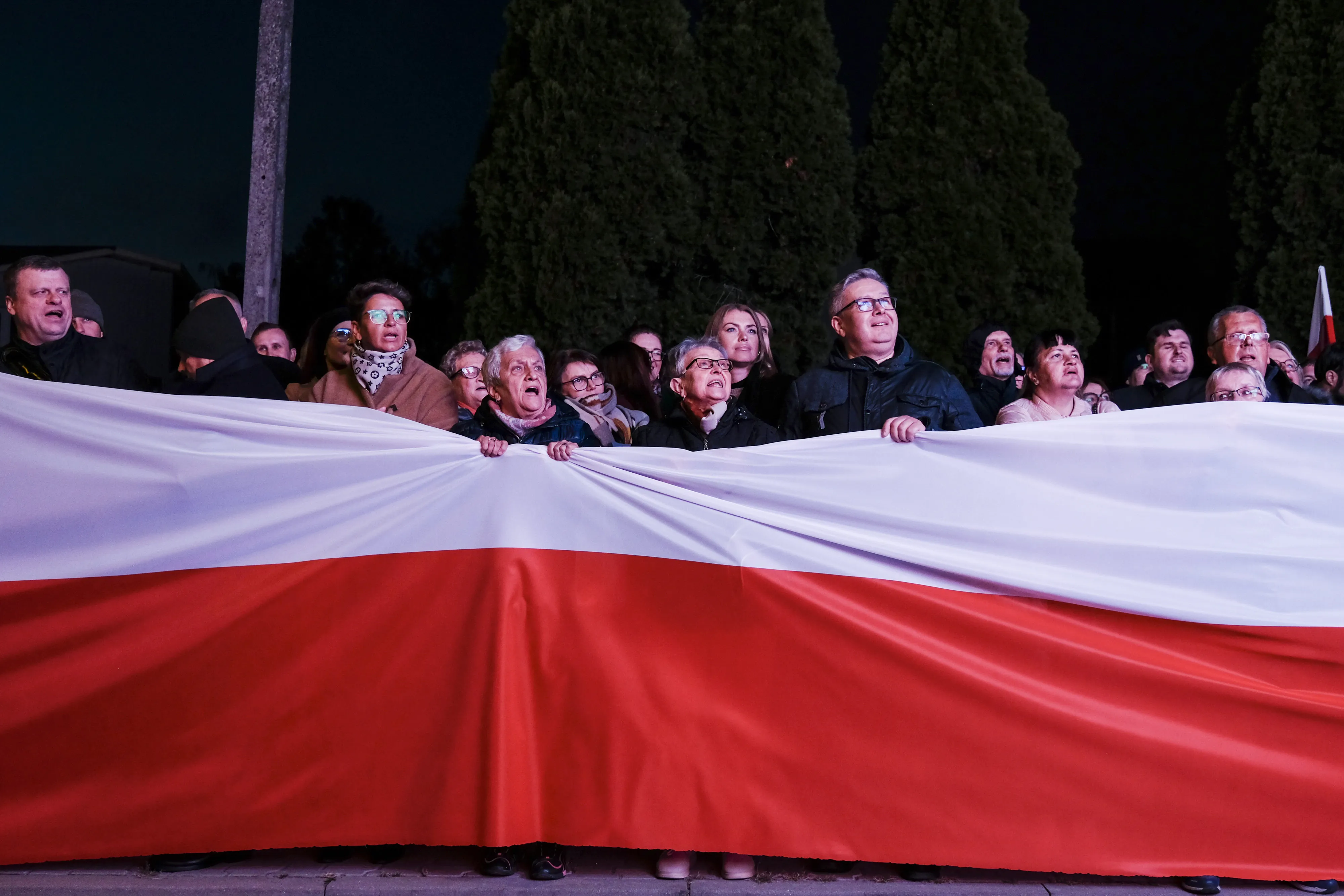 Law and Justice party supporters holding a Polish national flag banner watch an election debate on a screen outside the ATM studio in Warsaw, Poland, on&nbsp;Oct. 9.