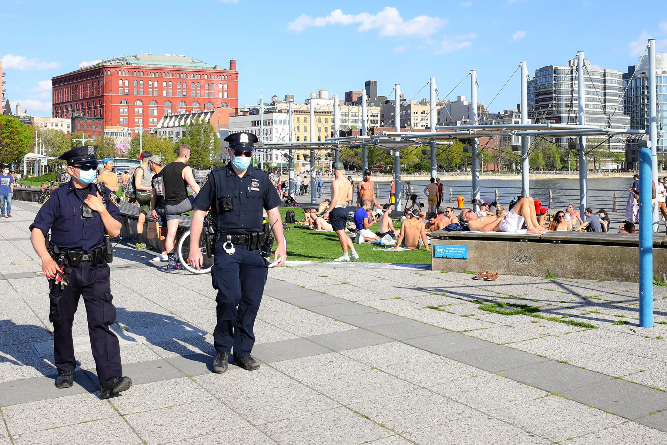 New York City police officers wearing protective masks walk past people sitting on a grassy area&nbsp;around the Hudson River on May 3.