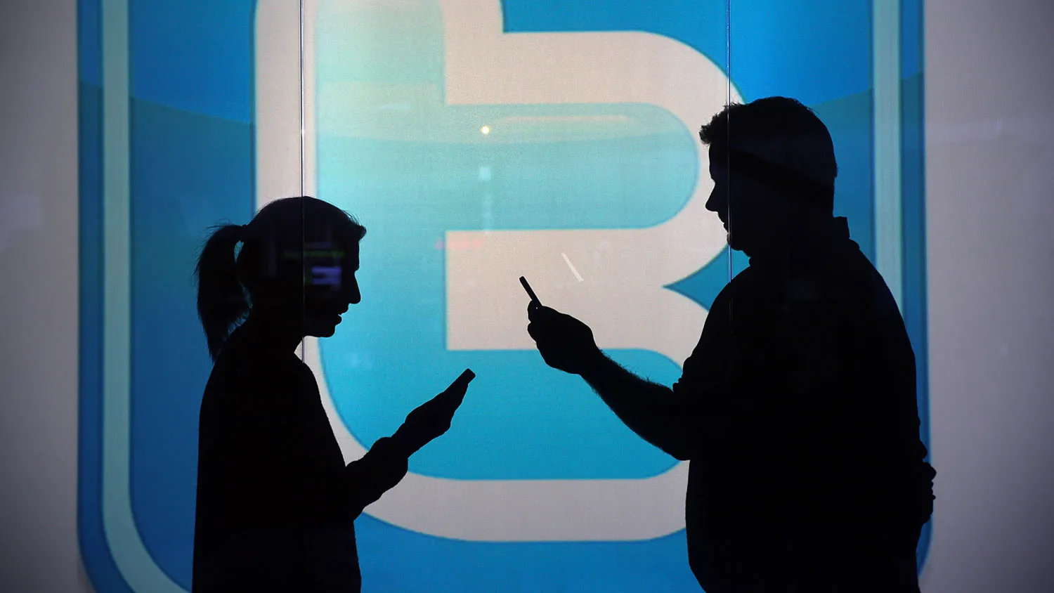 People are seen as silhouettes as they check mobile devices whilst standing against an illuminated wall bearing Twitter Inc.'s logo in this arranged photograph in London on Jan. 5, 2016.

