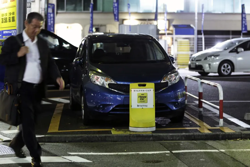 A pedestrian walks past a Nissan Motor Co. vehicle parked behind a sign for the “Times Car Plus” car sharing service at a car sharing station, operated by Park24 Co., in Tokyo, Japan.