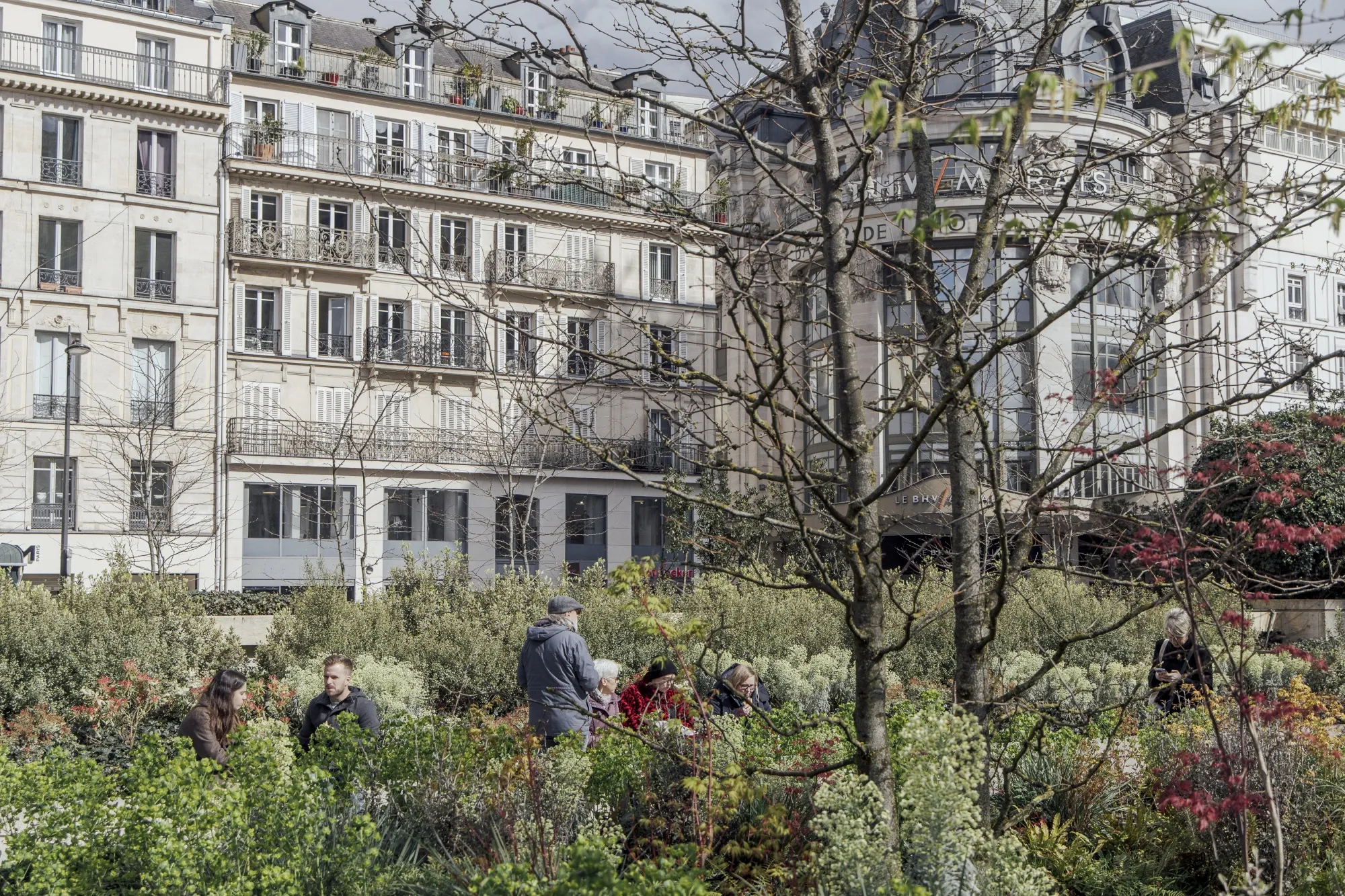 An urban garden filled with green bushes and small trees, with classic Haussmann-style buildings in the background. Several people are seen enjoying the space, sitting on benches or standing among the greenery. To the right, the ornate facade of the BHV Marais department store is visible through the branches of a budding tree.