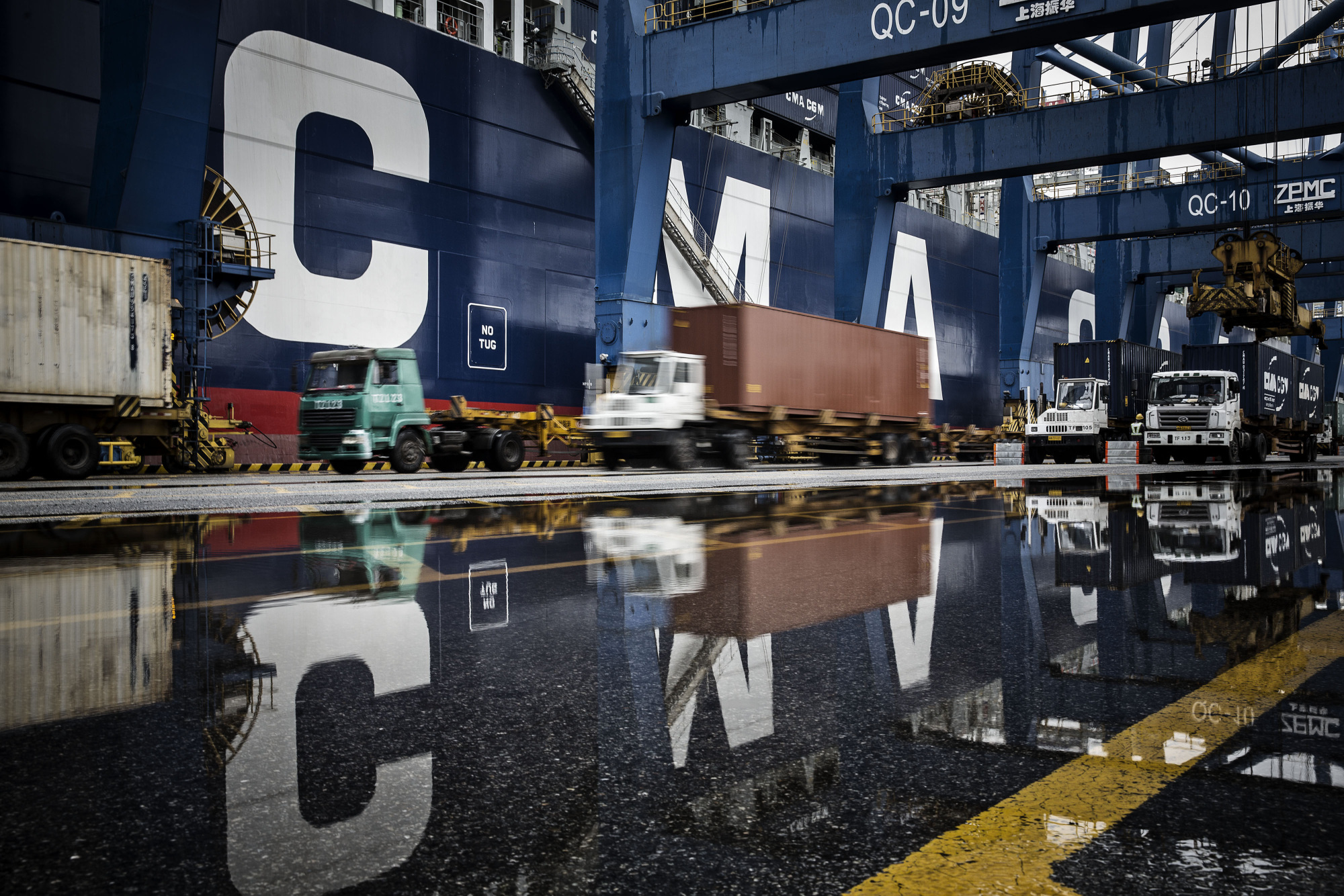 Trucks are reflected in a puddle as they wait in line to unload their containers onto a container ship docked at the Guangzhou Nansha Container Port in Guangzhou, China.