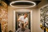 A blond middle-aged man is facing the camera and cutting a blond customer's hair, still wet from a shampoo. They're both in the center of the picture, the customer's back is facing the camera and their face cannot be seen. There is a large halo lamp hanging from the ceiling, and the wall on the left is of exposed brick. There is the painting of the tree on the wall to the right of Metzler, and behind him is a large and fancy mirror.